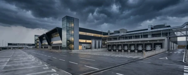 Vista amplia de una planta industrial moderna bajo un cielo oscuro cargado de nubes de tormenta, con zona de carga exterior vacía en primer plano