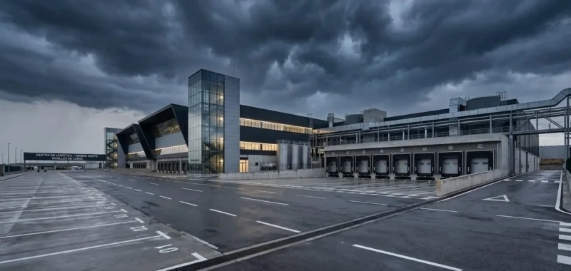 Vista amplia de una planta industrial moderna bajo un cielo oscuro cargado de nubes de tormenta, con zona de carga exterior vacía en primer plano